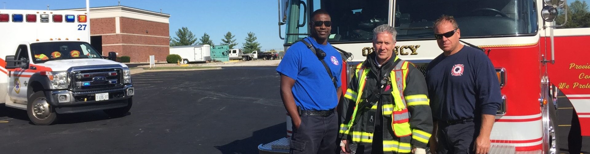 A police officer, firefighter, and medic pose in front of emergency vehicles