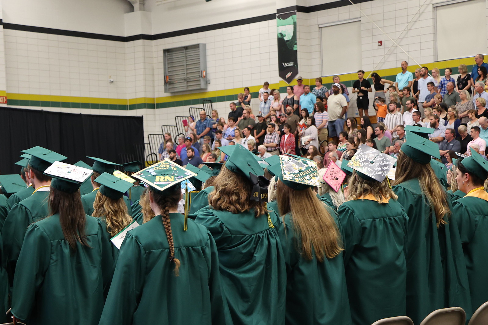 JWCC graduates stand in front of their chairs in green graduation caps and gowns
