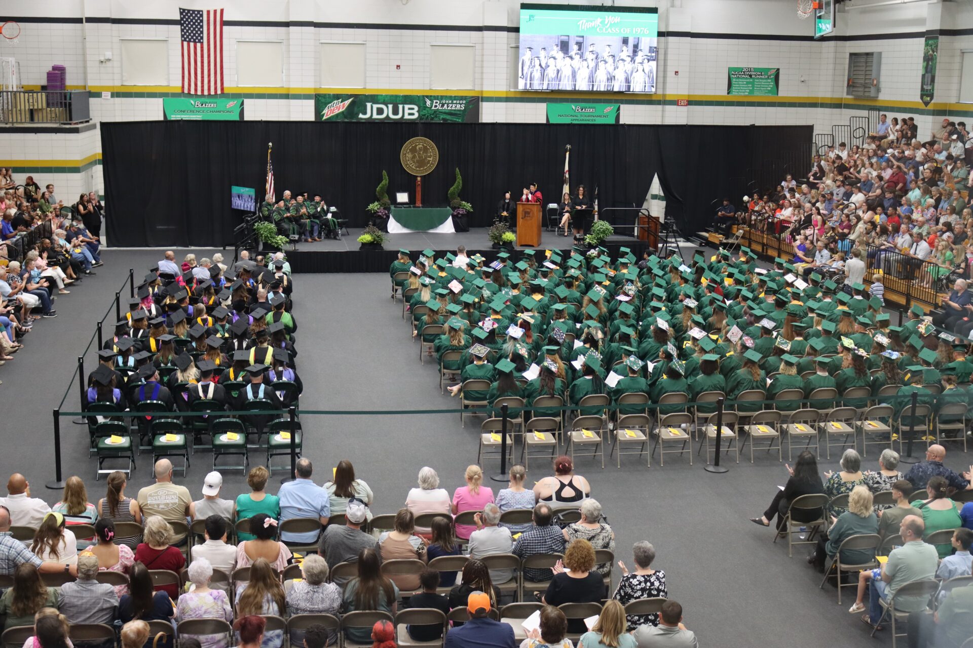 The JWCC Class of 2025 listens to commencement speakers during the graduation ceremony, as faculty, staff, and guests look on in a packed gymnasium. A tribute to the Class of 1976 is displayed on the screen above the stage.