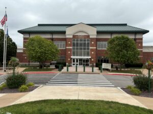 Front entrance of the John Wood Community College Student Administrative Center, featuring a brick facade, large glass entryway, landscaped grounds, and a pedestrian crosswalk leading to the building.